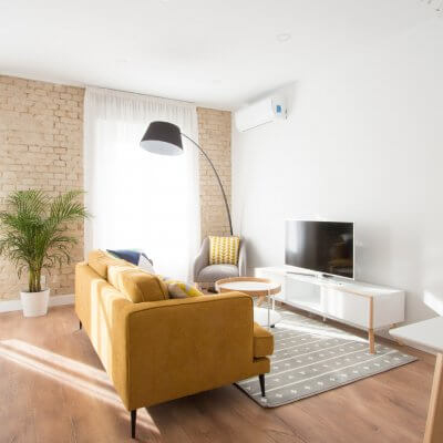 Proyecto de reforma de vivienda en Chamberí II (Madrid) de R de Room. Zona de estar con sofá modelo MAD en color mostaza de estilo mid-century, mueble de tv y mesa de centro con ruedas de estilo escandinavo. Butaca gris y lámpara de pie arco en color negro. Pared de ladrillo visto original.
