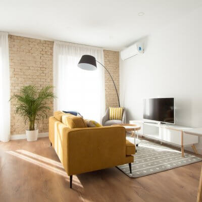 Proyecto de reforma de vivienda en Chamberí II (Madrid) de R de Room. Zona de estar con sofá modelo MAD en color mostaza de estilo mid-century, mueble de tv y mesa de centro con ruedas de estilo escandinavo. Butaca gris y lámpara de pie arco en color negro. Pared de ladrillo visto original.