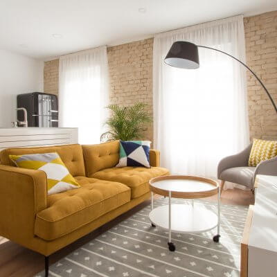 Proyecto de reforma de vivienda en Chamberí II (Madrid) de R de Room. Zona de estar con sofá modelo MAD en color mostaza de estilo mid-century, mueble de tv y mesa de centro con ruedas de estilo escandinavo. Butaca gris y lámpara de pie arco en color negro. Pared de ladrillo visto original. Cocina abierta con península y frigorífico SMEG. Radiador Runtal.