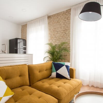 Proyecto de reforma de vivienda en Chamberí II (Madrid) de R de Room. Zona de estar con sofá modelo MAD en color mostaza de estilo mid-century, mueble de tv y mesa de centro con ruedas de estilo escandinavo. Butaca gris y lámpara de pie arco en color negro. Pared de ladrillo visto original. Cocina abierta con península y frigorífico SMEG. Radiador Runtal.