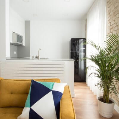 Proyecto de reforma de vivienda en Chamberí II (Madrid) de R de Room. Cocina abierta con península y frigorífico SMEG en color negro. Sofá modelo MAD en color mostaza de estilo mid-century. Pared de ladrillo visto original. Radiador Runtal.