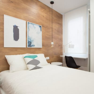 Proyecto de reforma de vivienda en Chamberí II (Madrid) de R de Room. Dormitorio principal con suelo laminado de madera y pared del cabecero forrada en laminado de madera. Lámparas colgantes de mármol blanco bombilla Edison. Mesita de noche modelo BLOCK en color blanco. Zona de escritorio.