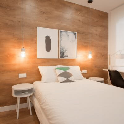 Proyecto de reforma de vivienda en Chamberí II (Madrid) de R de Room. Dormitorio principal con suelo laminado de madera y pared del cabecero forrada en laminado de madera. Lámparas colgantes de mármol blanco bombilla Edison. Mesita de noche modelo BLOCK en color blanco. Zona de escritorio.