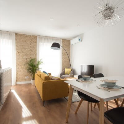 Proyecto de reforma de vivienda en Chamberí II (Madrid) de R de Room. Estar-comedor de estilo escandinavo. Lámpara de techo Sputnik resturada. Sofá modelo MAD en color mostaza y muebles de diseño nórdico en blanco y madera. Láminas de gran formado de Desenio.