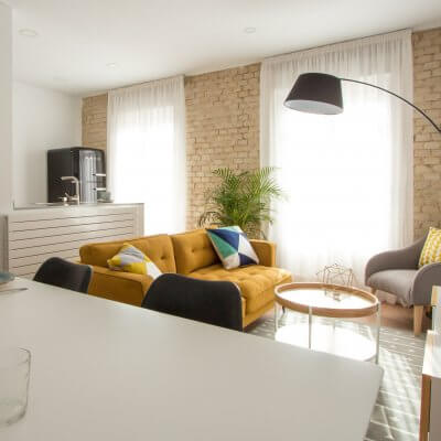 Proyecto de reforma de vivienda en Chamberí II (Madrid) de R de Room. Estar-comedor de estilo escandinavo. Lámpara de techo Sputnik resturada. Sofá modelo MAD en color mostaza y muebles de diseño nórdico en blanco y madera. Láminas de gran formado de Desenio.