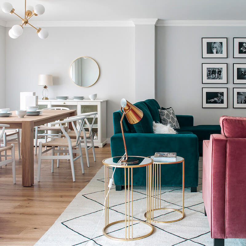 Salón y comedor de vivienda en Madrid. Proyecto de reforma e interiorismo de R de Room. Sofá tapizado en terciopelo verde y butaca tapizada en terciopelo rosa. Mesillas auxiliares doradas y alfombra tipo Beni Ouarain, blanca con rombos negros. Mesa de madera y lámpara suspendida dorada. Collage de cuadros.