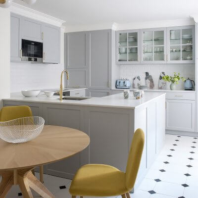 Cocina con península en Madrid. Office con mesa circular de madera de roble y sillas tapizadas en tela de color mostaza. Suelo blanco con rombos negros. Encimera de mármol Calacatta. Cocina panelada con puertas plafonadas. Cocina gris con tiradores dorados. Proyecto de reforma e interiorismo de R de Room.