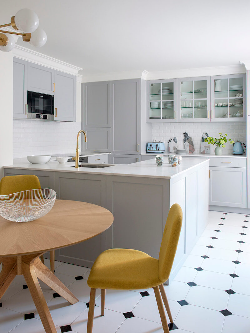 Cocina con península en Madrid. Office con mesa circular de madera de roble y sillas tapizadas en tela de color mostaza. Suelo blanco con rombos negros. Encimera de mármol Calacatta. Cocina panelada con puertas plafonadas. Cocina gris con tiradores dorados. Proyecto de reforma e interiorismo de R de Room.