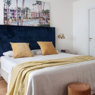 Dormitorio con cabecero tapizado en terciopelo azul con capitoné. Cojines y complementos mostaza. Dormitorio blanco y luminoso con toques de color. Apliques de pared dorados. Proyecto de reforma e interiorismo de R de Room.
