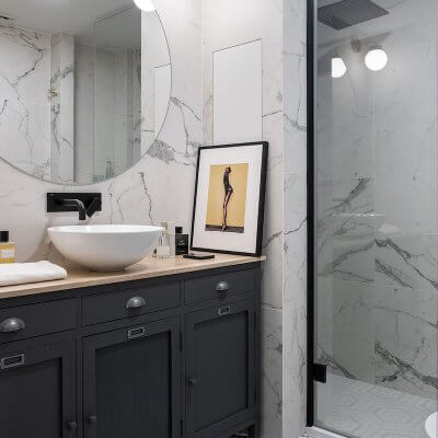 Baño en suite de dormitorio en Madrid. Aparador adaptado como mueble de baño. Grifería negra, mampara negra y apliques dorados. Porcelánico efecto mármol Calacatta. Espejo circular. Proyecto de reforma e interiorismo de R de Room.