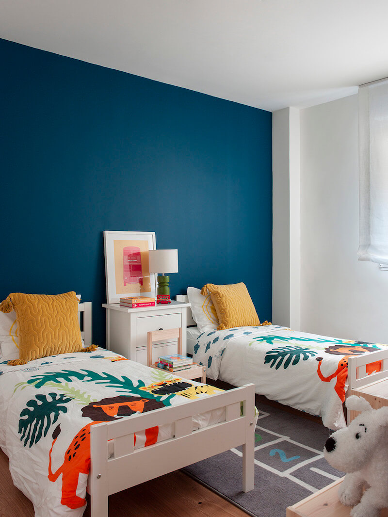 Dormitorio infantil de vivienda en Madrid. Pared del cabecero pintada en color azul pato. Colchas con estampado de animales y cojines mostaza. Cuarto de niños. Proyecto de reforma e interiorismo de R de Room.