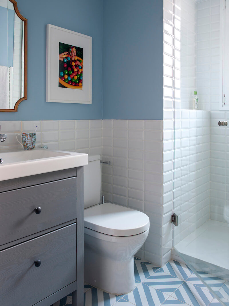 Baño con ducha en Madrid. Zócalo de azulejo metro blanco y pared pintada en color azul. Baldosa con estampado blanco y azul. Puerta blanca plafonada con tiradores dorados. Mueble de baño Hemnes de IKEA. Proyecto de reforma e interiorismo de R de Room.