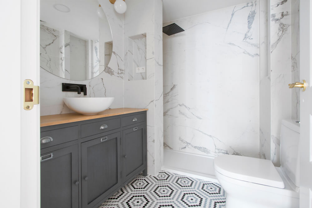 Baño en suite de dormitorio en Madrid. Aparador adaptado como mueble de baño. Grifería negra, mampara negra y apliques dorados. Porcelánico efecto mármol Calacatta. Espejo circular. Proyecto de reforma e interiorismo de R de Room.