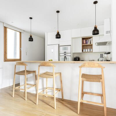 Barra en cocina abierta. Predomina el blanco con toques de madera. La encimera y los taburetes son el toque cálido perfecto para esta casa. Proyecto de R de Room.
