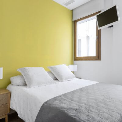 Sencillo pero efectivo dormitorio para invitados. Mesitas de madera, ropa de cama neutra y pared pintada en amarillo. Proyecto de R de Room.