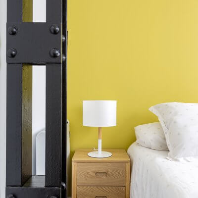 Sencillo pero efectivo dormitorio para invitados. Mesitas de madera, ropa de cama neutra y pared pintada en amarillo. Proyecto de R de Room.
