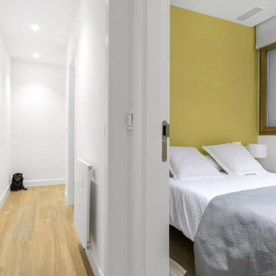 Sencillo pero efectivo dormitorio para invitados. Mesitas de madera, ropa de cama neutra y pared pintada en amarillo. Vista desde el pasillo. Proyecto de R de Room.